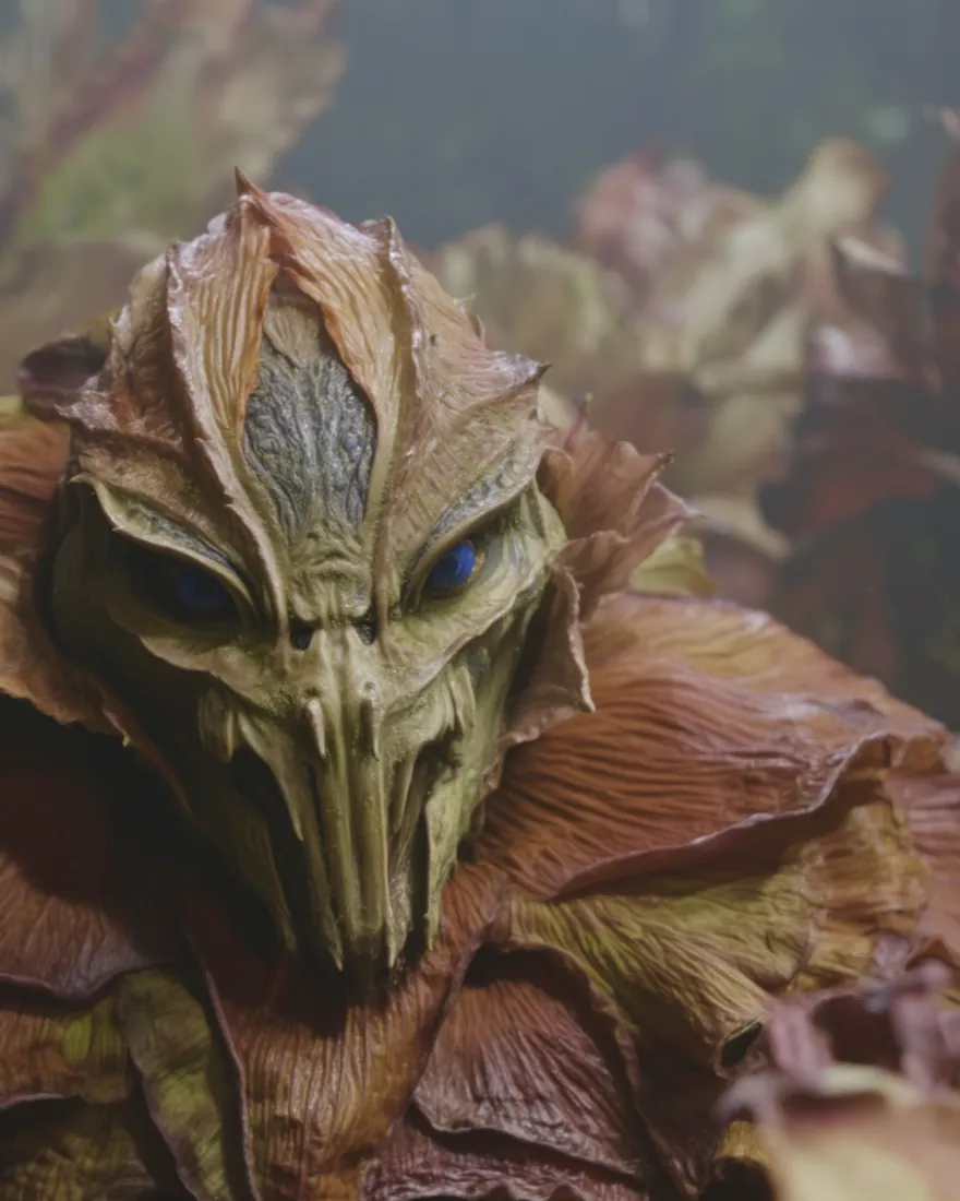 A Foigan among the leaves, the face insectoid beneath folded and puffed petals, the two dark blue eyes steady.